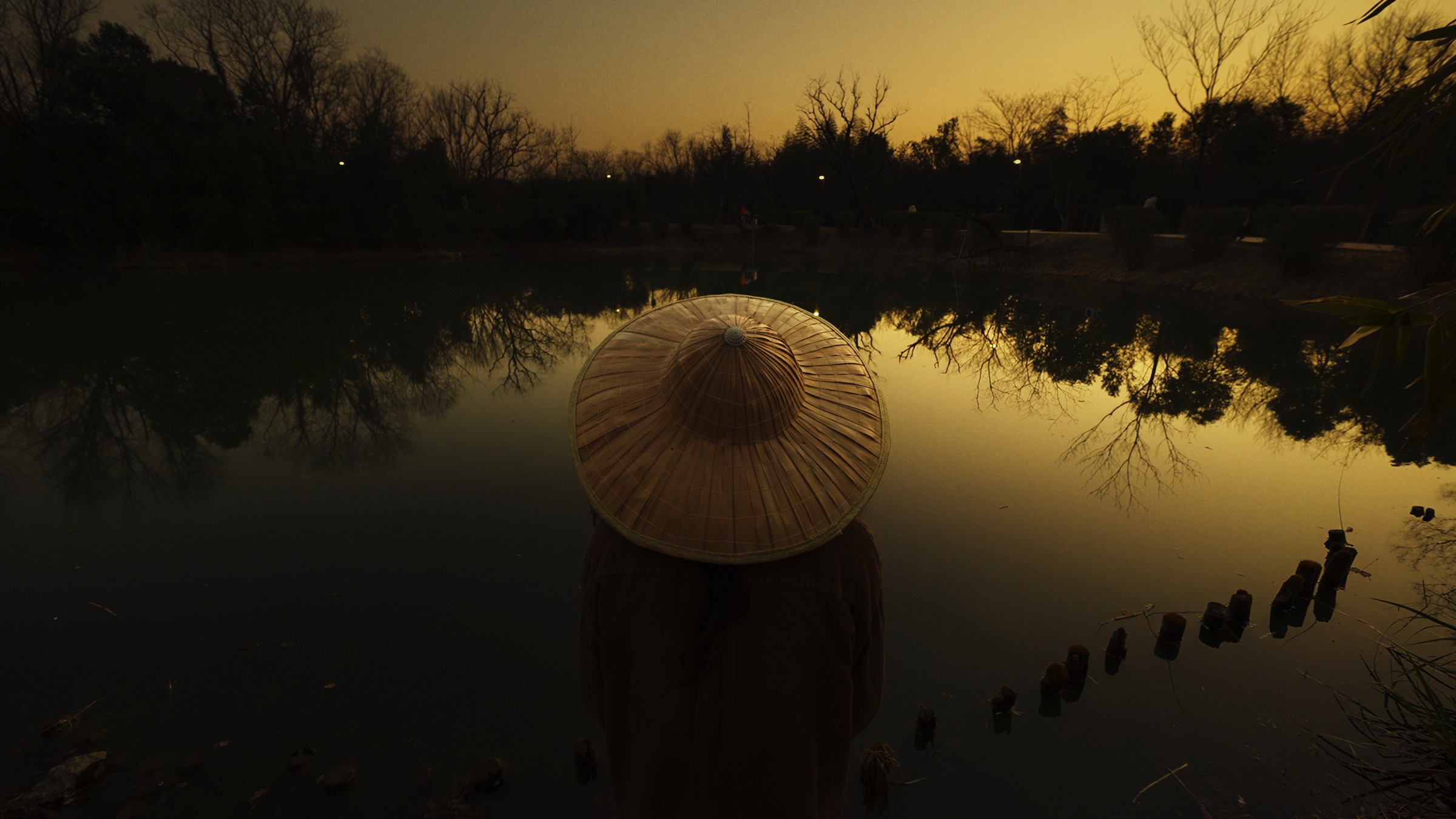 Figure in a wide-brimmed hat standing in a lake at dusk, before